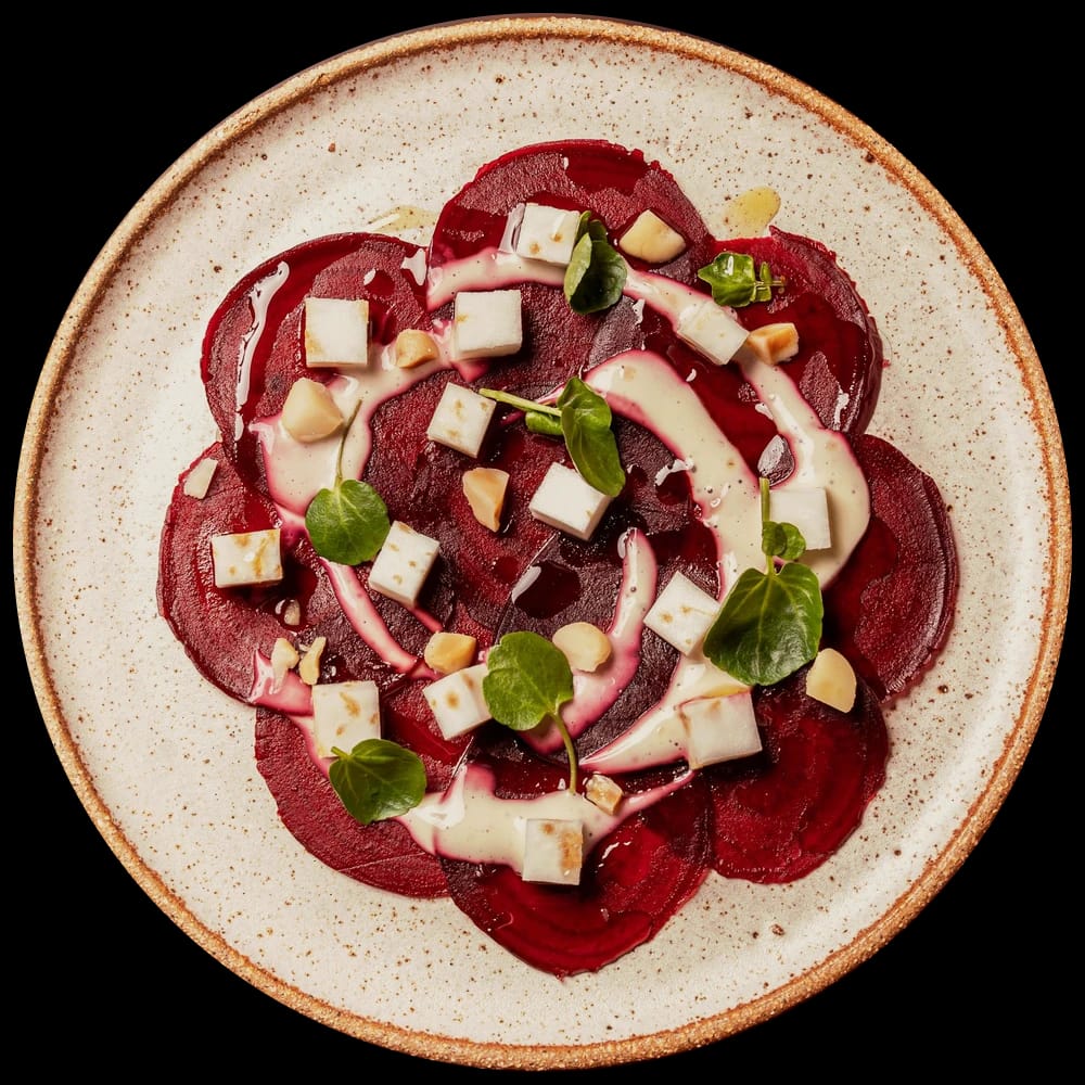 Carpaccio de beterraba em lâminas com cubos de queijo de cabra, amêndoas, fios de creme e folhas de capuchinha, num prato de cerâmica pontilhada visto de cima